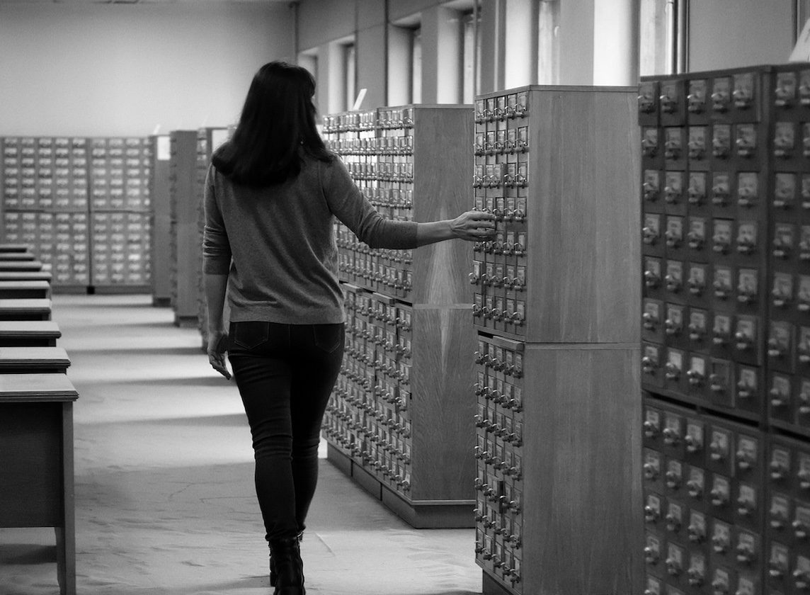 Woman walking along rows of old card catalogue drawers in an archive.