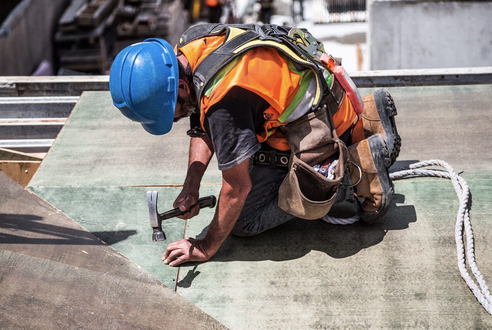 Builder in a hard hat and safety harness nailing on a roof.